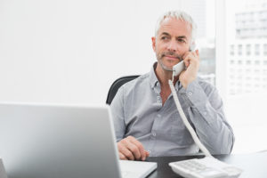 Serious mature businessman on call in front of laptop at desk in a bright office